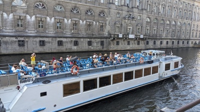 The bottom screen shows a blue-white boat with tourists on the deck whereas the background shows a grey government building