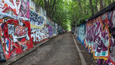 The focus point of a road ends in green trees but on the left and right side are walls covered in colorful graffiti