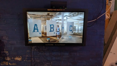 A screenshot of a video is photographed with Vladimir standing in front of canvases showing his works