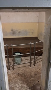 A prison cell is shown with a wooden surface in the back and a bucket in front